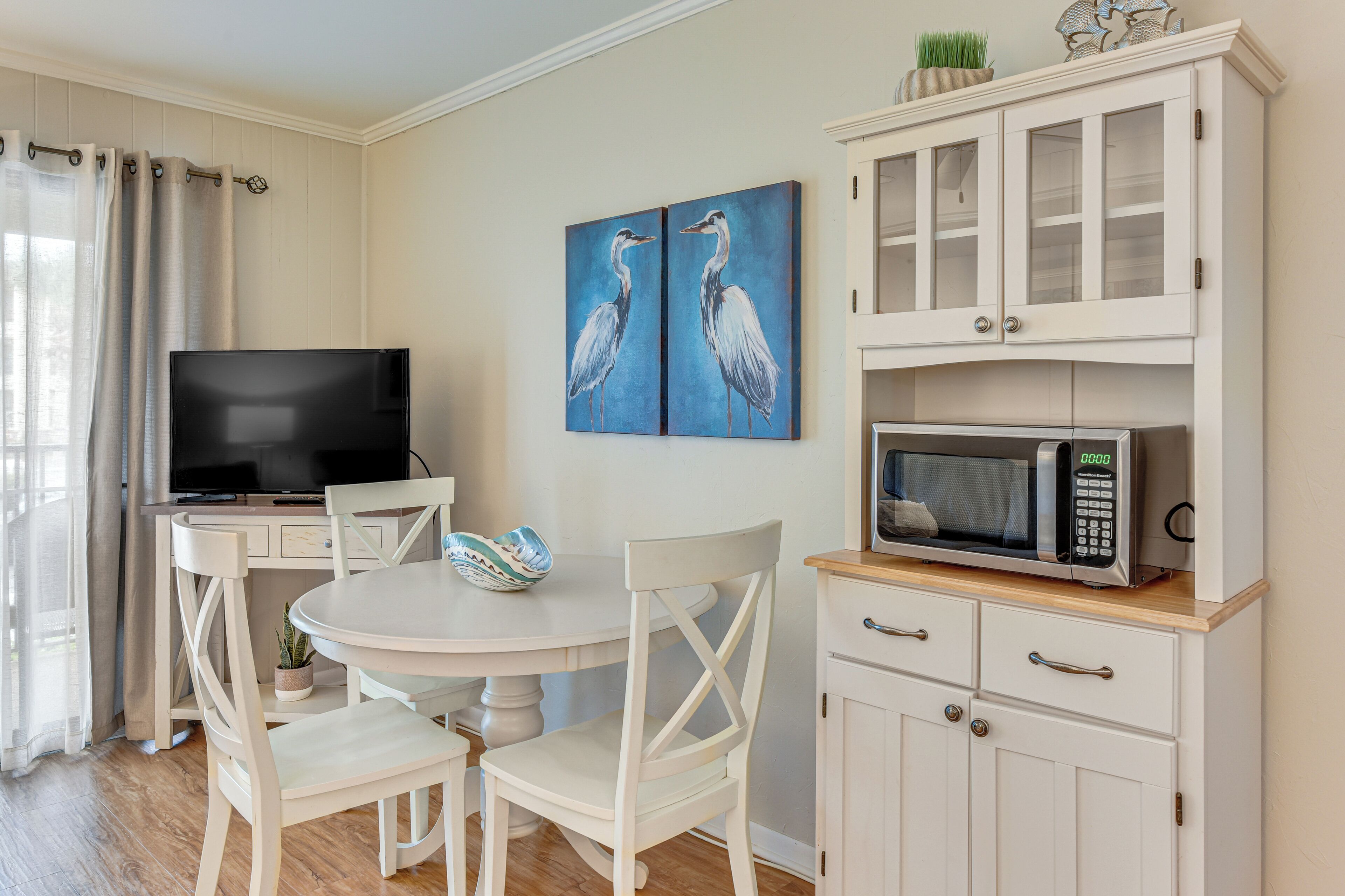 A dining area with a round table, white chairs, a television, a cabinet with a microwave, and a painting of birds on the wall.
