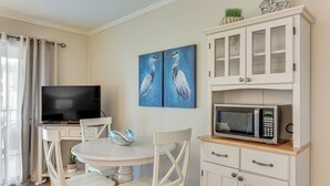 Un coin repas avec une table ronde, des chaises blanches, un téléviseur, un meuble contenant un four à micro-ondes et un tableau représentant des oiseaux accroché au mur.