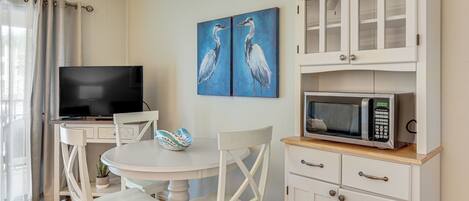 A dining area with a round table, white chairs, a television, a cabinet with a microwave, and a painting of birds on the wall.