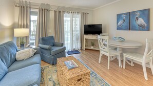 Un salon comprenant un canapé, un fauteuil, une table basse, un téléviseur, une table à manger avec des chaises, et des œuvres d’art accrochées au mur.