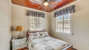 A bedroom with a wooden ceiling, a bed with a quilt, a nightstand, a lamp, and two windows with blinds. - Paddle, Fish & Swim! Hernando Lake Cabin (Hernando)