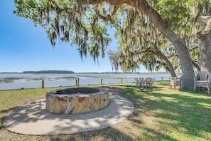A stone fire pit surrounded by a concrete circle, outdoor furniture, and a view of a lake with a distant shoreline.