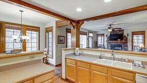 A kitchen with wooden cabinets, a large island with a sink, a stone fireplace, and a ceiling fan. - Forest-view Balcony & Trail Access! Burnsville Gem (Burnsville)