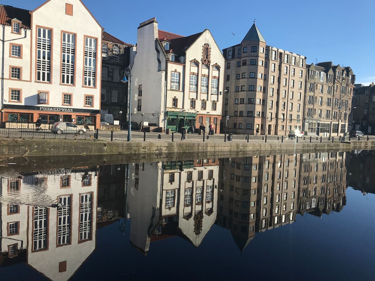 A waterfront with buildings reflecting in calm water, including a visible Pizza Express restaurant.