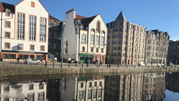 Een waterkant met gebouwen die weerspiegelen in kalm water, waaronder een zichtbare Pizza Express restaurant.