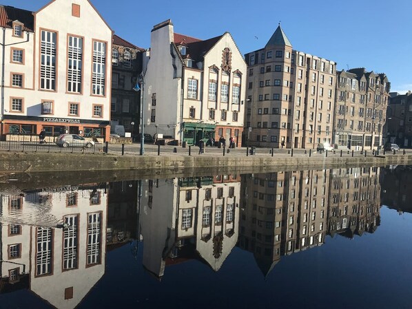 Ein Ufer mit Gebäuden, die sich in ruhigem Wasser spiegeln, darunter ein sichtbar gelegenes Pizza Express Restaurant.