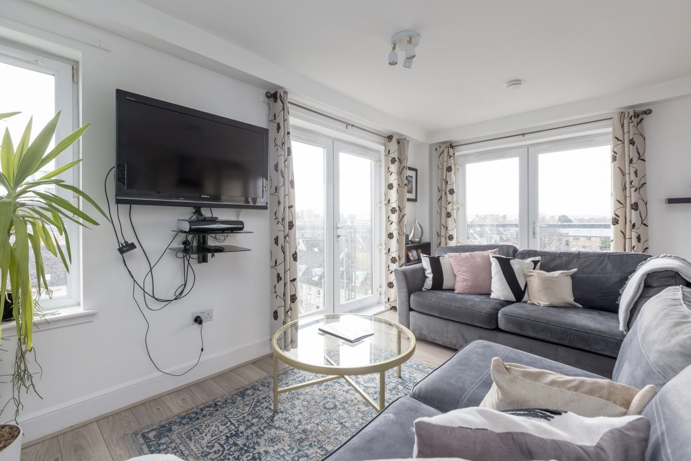 A modern living room with a grey sofa, a glass coffee table, and a flat-screen TV mounted on the wall.