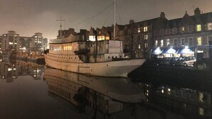 Een groot, meerdekker schip afgemeerd in een haven, met gebouwen op de achtergrond.