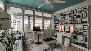Ein Zimmer mit Deckenventilator, Schreibtisch mit Computer, Bücherregal mit gerahmten Bildern und Blick durch große Fenster.