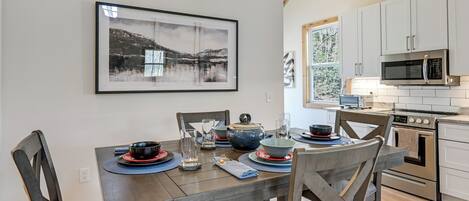 A dining table set with plates, bowls, glasses, and cutlery, with a painting above and a kitchen in the background.