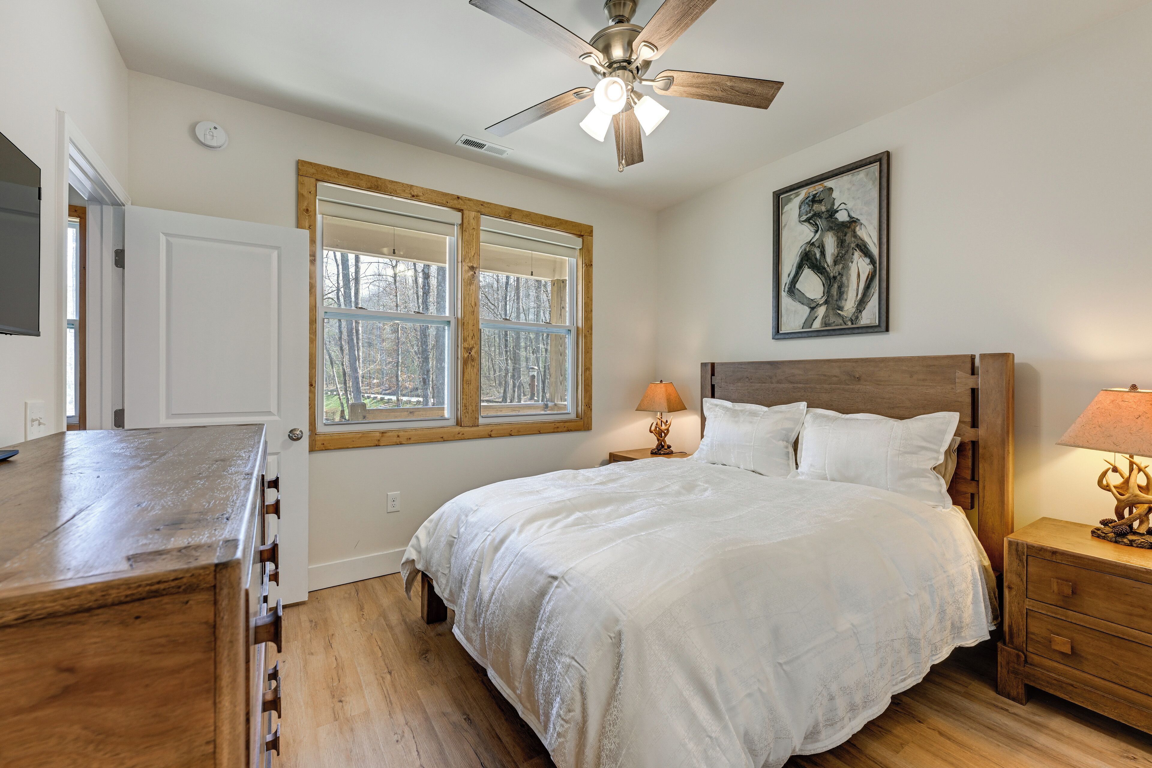 A bedroom with a bed, a ceiling fan, a window, a nightstand, and a wall-mounted artwork.