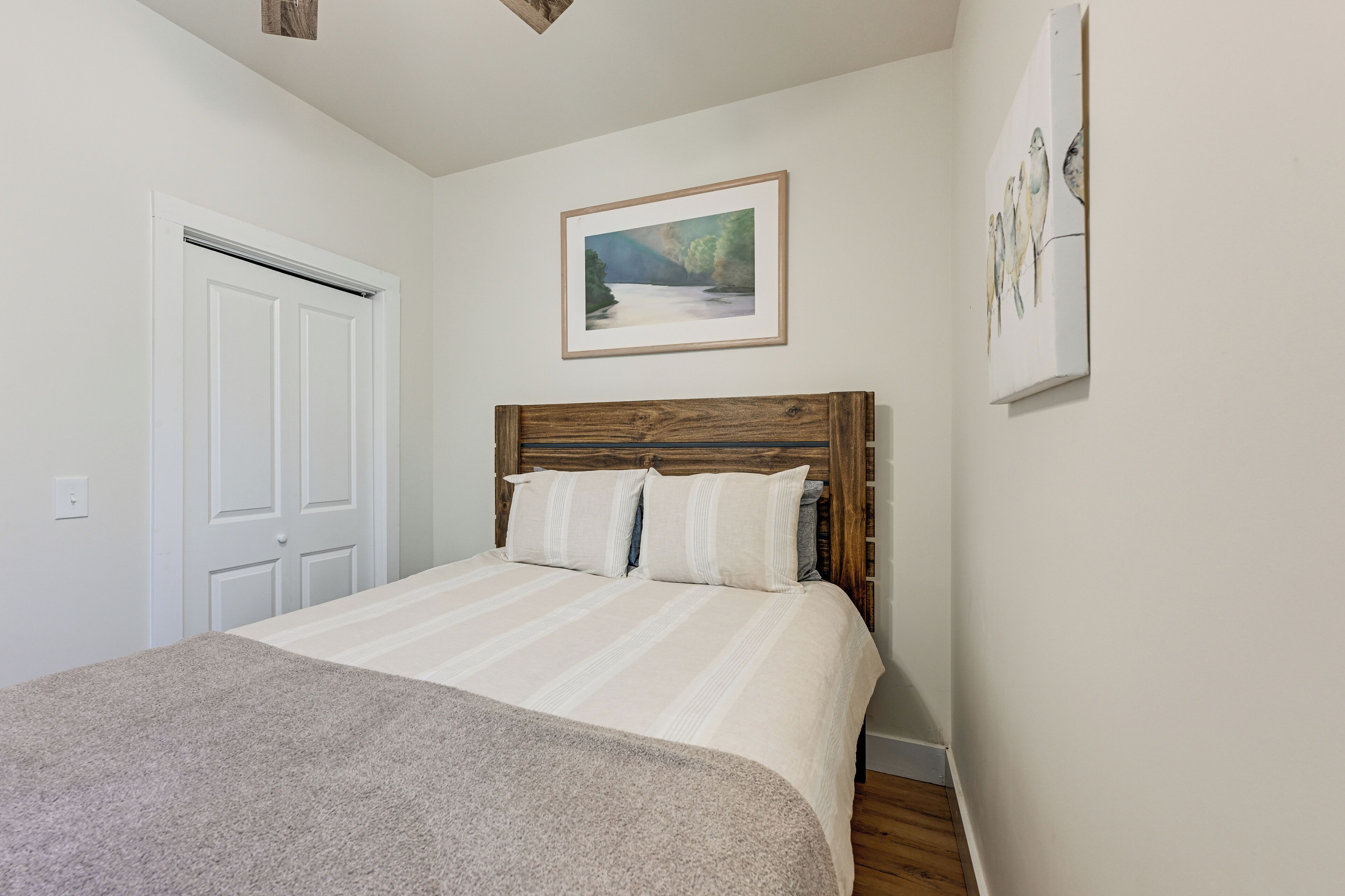 A bedroom with a wooden headboard, a bed with white bedding, a framed landscape painting, and a white door.