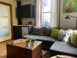 Ein modernes Wohnzimmer mit einer Couch, einem Couchtisch mit Büchern und Pflanzen, einer Küchenzeile mit Schränken und einem Fenster mit Vorhängen.
