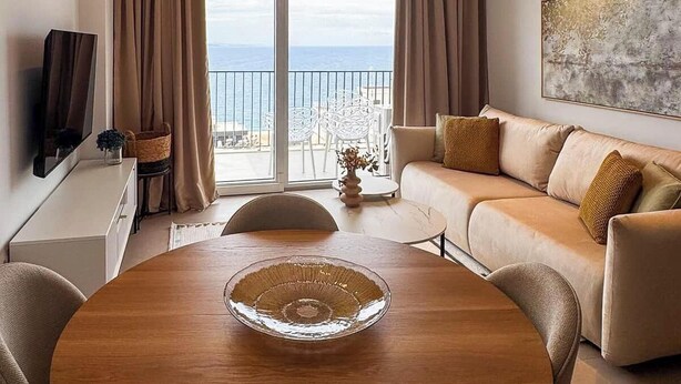 Una sala de estar moderna con una mesa redonda de madera, un sofá, un televisor y un ventanal con vistas al mar.