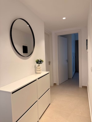 A hallway with a white wall, a round mirror, a white dresser with drawers, and a potted plant. - Arletta - Panoramic View (Sarandë)