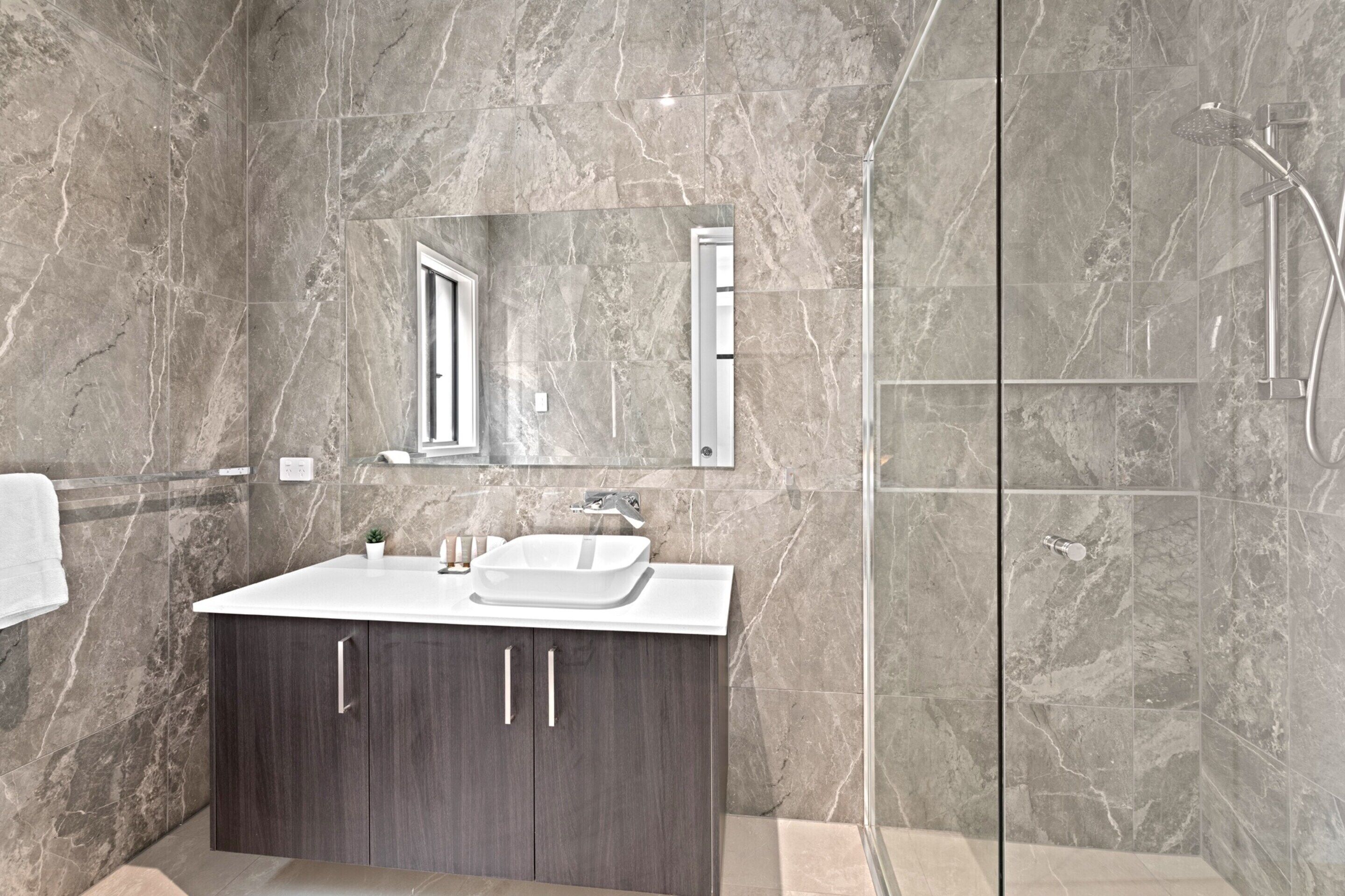 A modern bathroom with a marble wall, a glass shower enclosure, a white sink, and dark wooden cabinets.