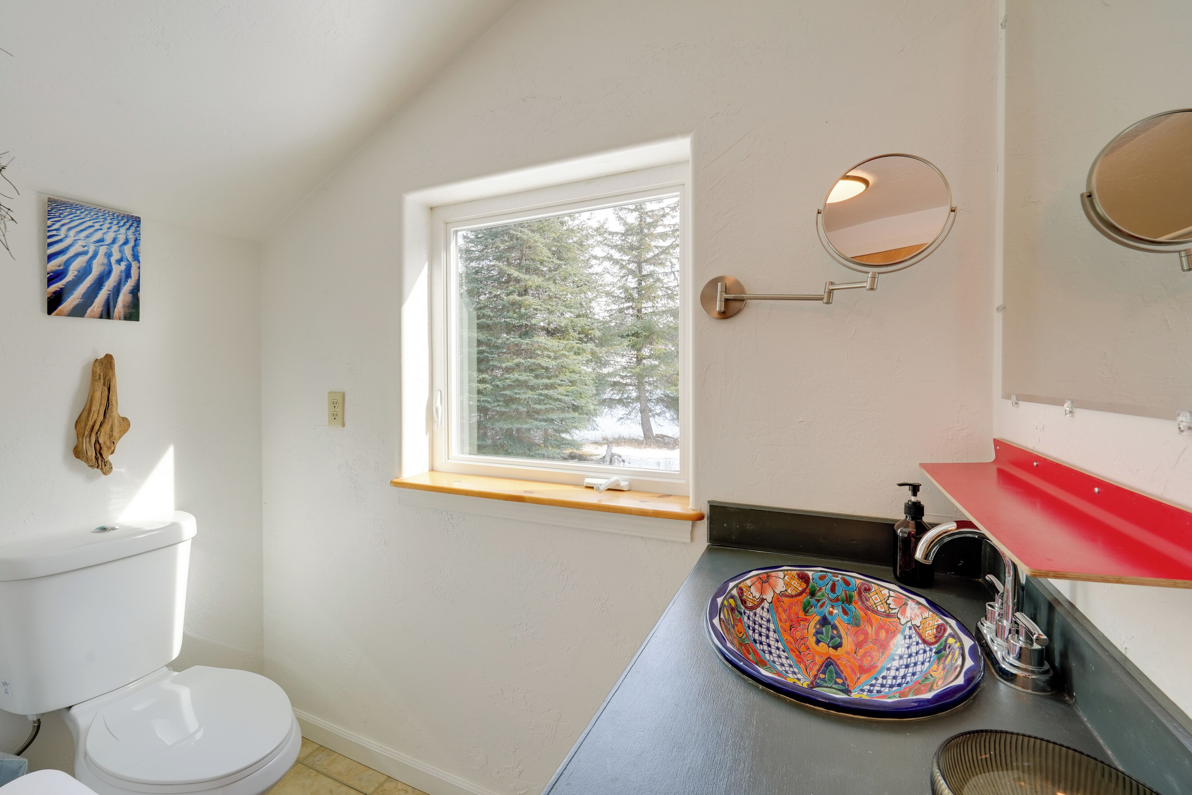 Une salle de bain avec des toilettes, une fenêtre, un lavabo avec un receveur coloré et un miroir.