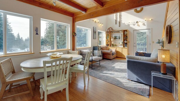 A spacious living area with a dining table, chairs, and a sofa. Large windows provide natural light and views of the outdoors. - Secluded Homer Escape With Deck in Quite Woods (Homer)