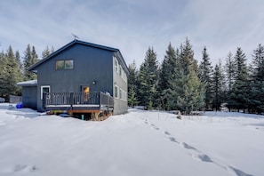A two-story house with a deck, surrounded by snow and evergreen trees.