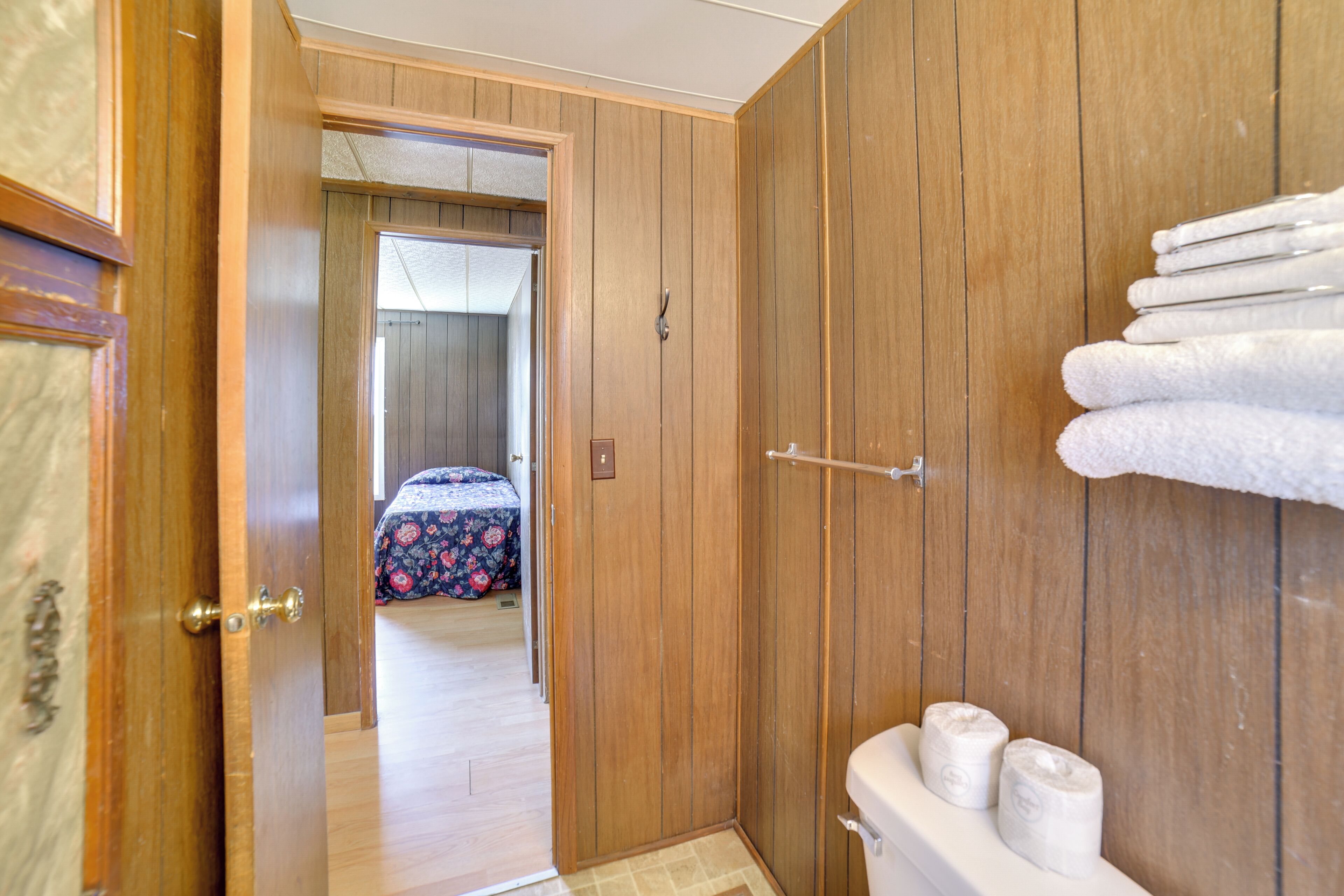 A wooden-paneled bathroom with a toilet, a towel rack, and a visible door leading to a bedroom.
