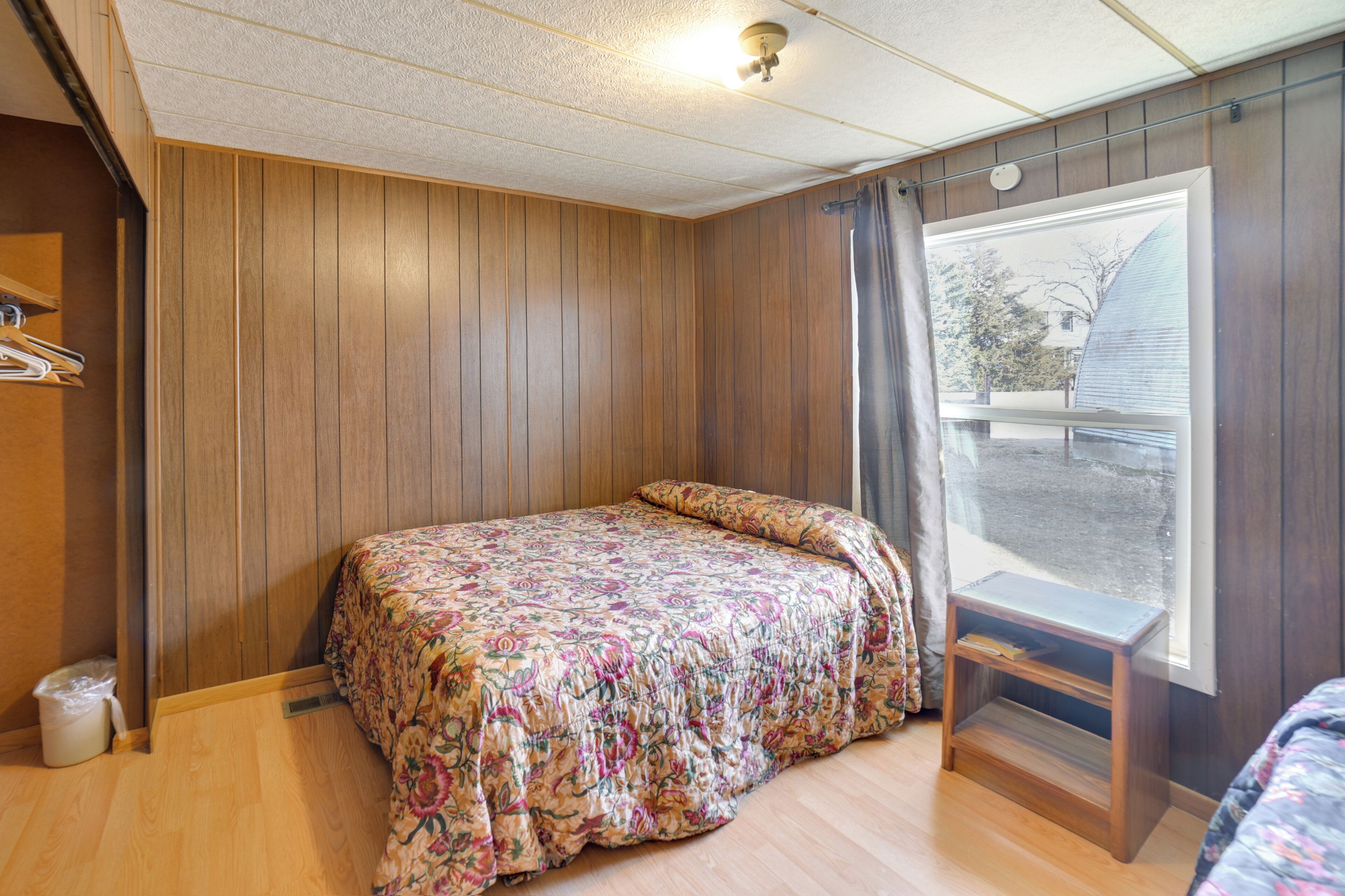 A room with a wooden paneled wall, a bed with a floral bedspread, a small wooden nightstand, and a window with a view of a house and trees.