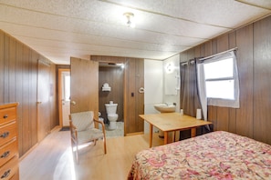 Un dormitorio con paredes de madera, una cama con colcha floral, un aparador de madera, una silla, una mesa de madera, una ventana con cortinas, un lavabo y un inodoro.