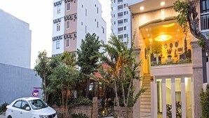 Entrada de hotel com um carro estacionado em frente, cercada por vegetação e um edifício com letreiro de neon.