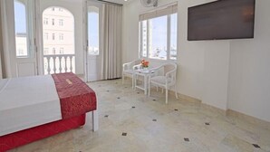 Quarto de hotel com uma cama, uma mesinha com cadeiras, televisão e janelas amplas.