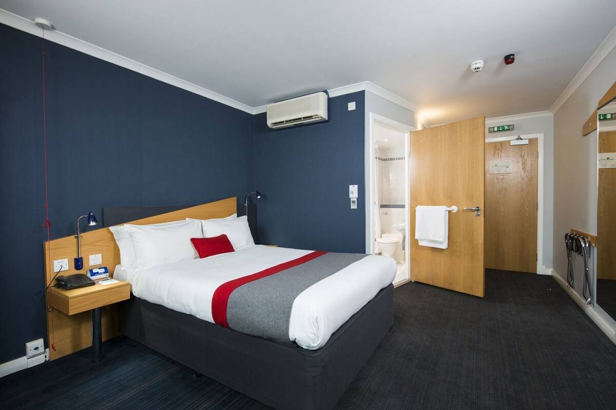 Habitación de hotel con cama, mesita de noche, lámpara, equipo de aire acondicionado y puerta que da a un baño.