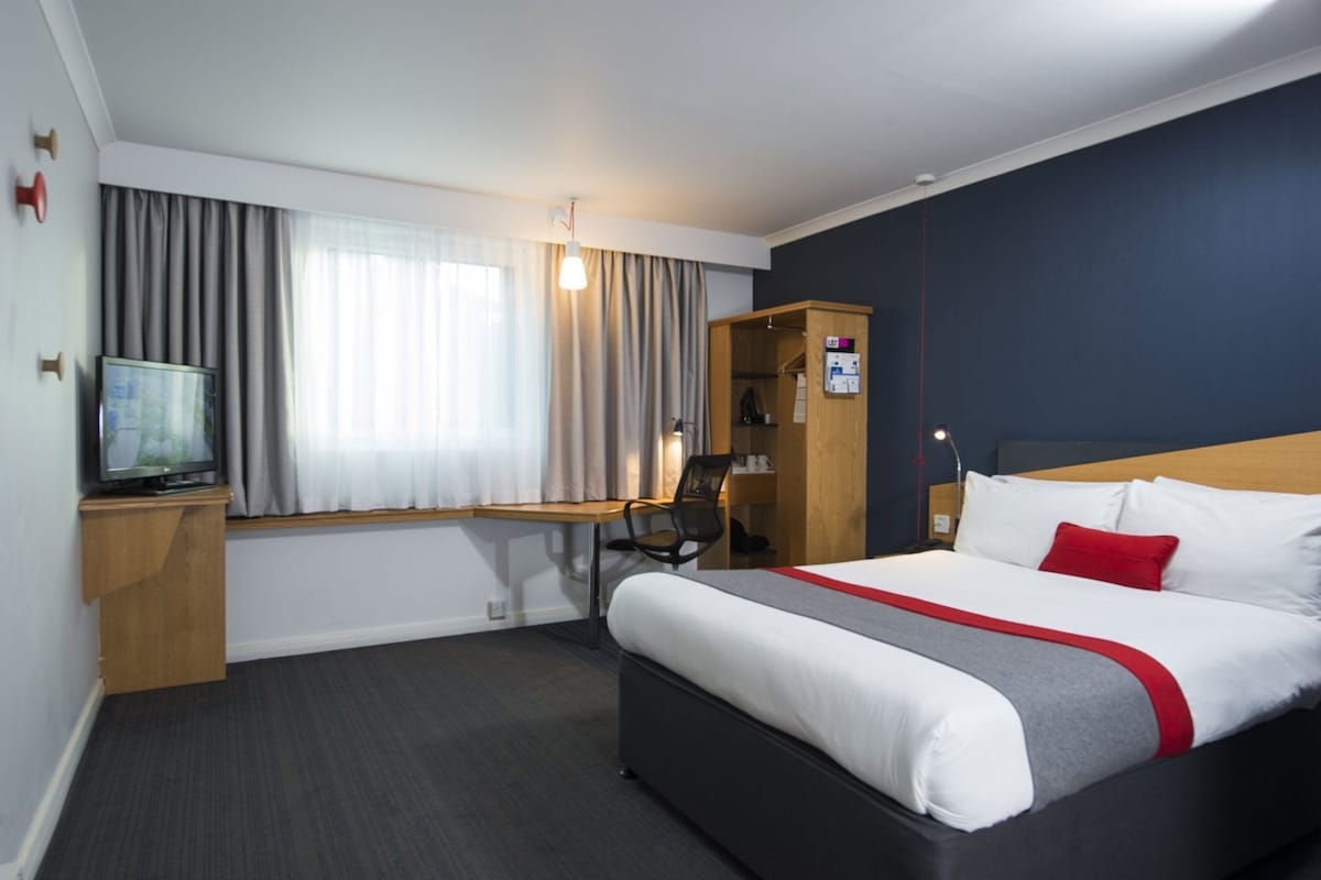 A hotel room with a bed, desk, chair, television, wardrobe, and a window with curtains.