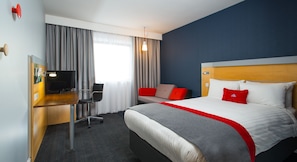 Quarto de hotel com cama, escrivaninha, cadeira, televisão e janela com cortinas.