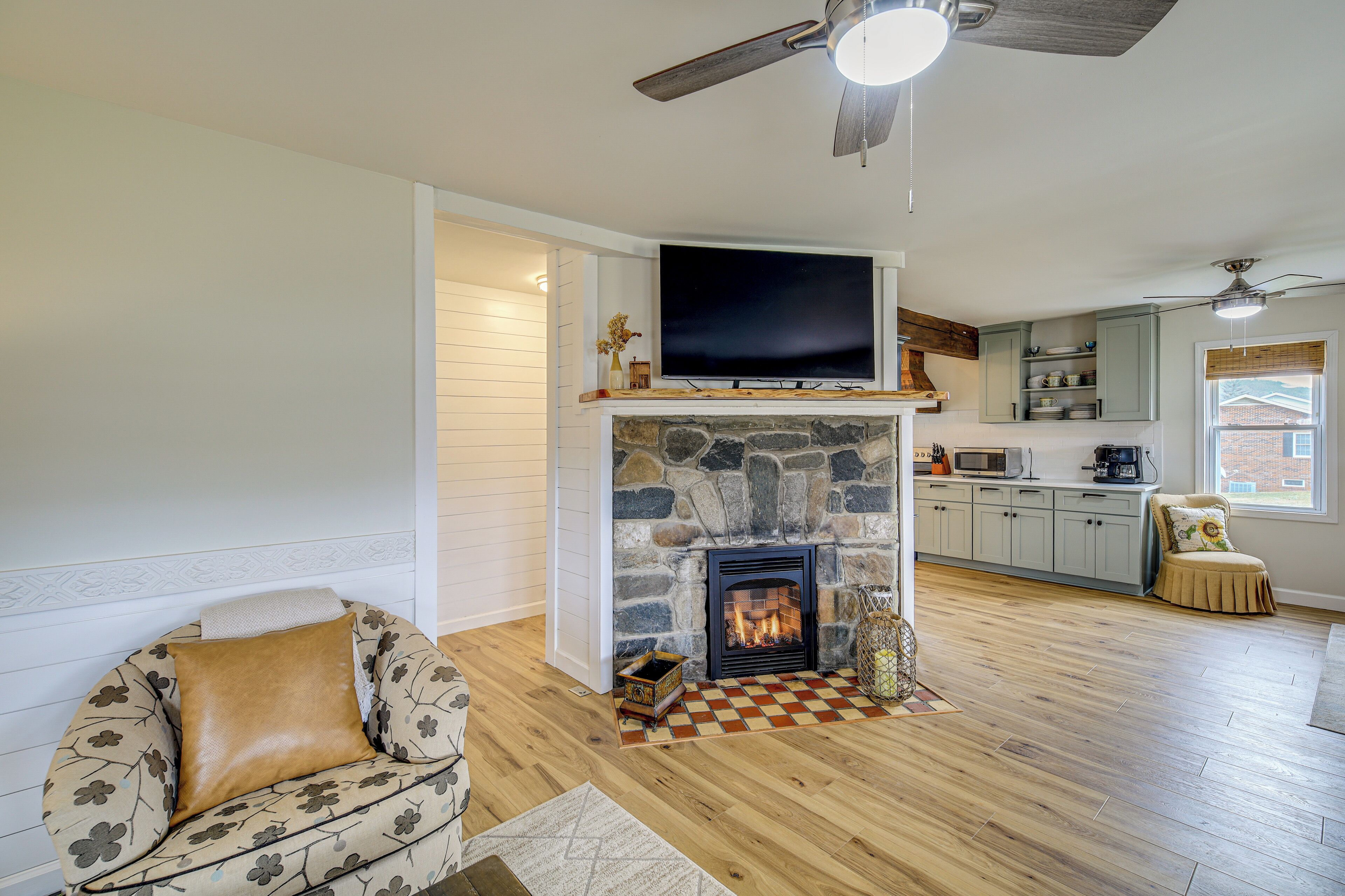 A living room with a stone fireplace, a television mounted above it, and a kitchen area in the background.