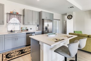 A modern kitchen with grey cabinets, a stainless steel refrigerator, a dining area with a white table, and a green sofa. - Unwind in Kissimmee! Rural Retreat for Families (Poinciana)