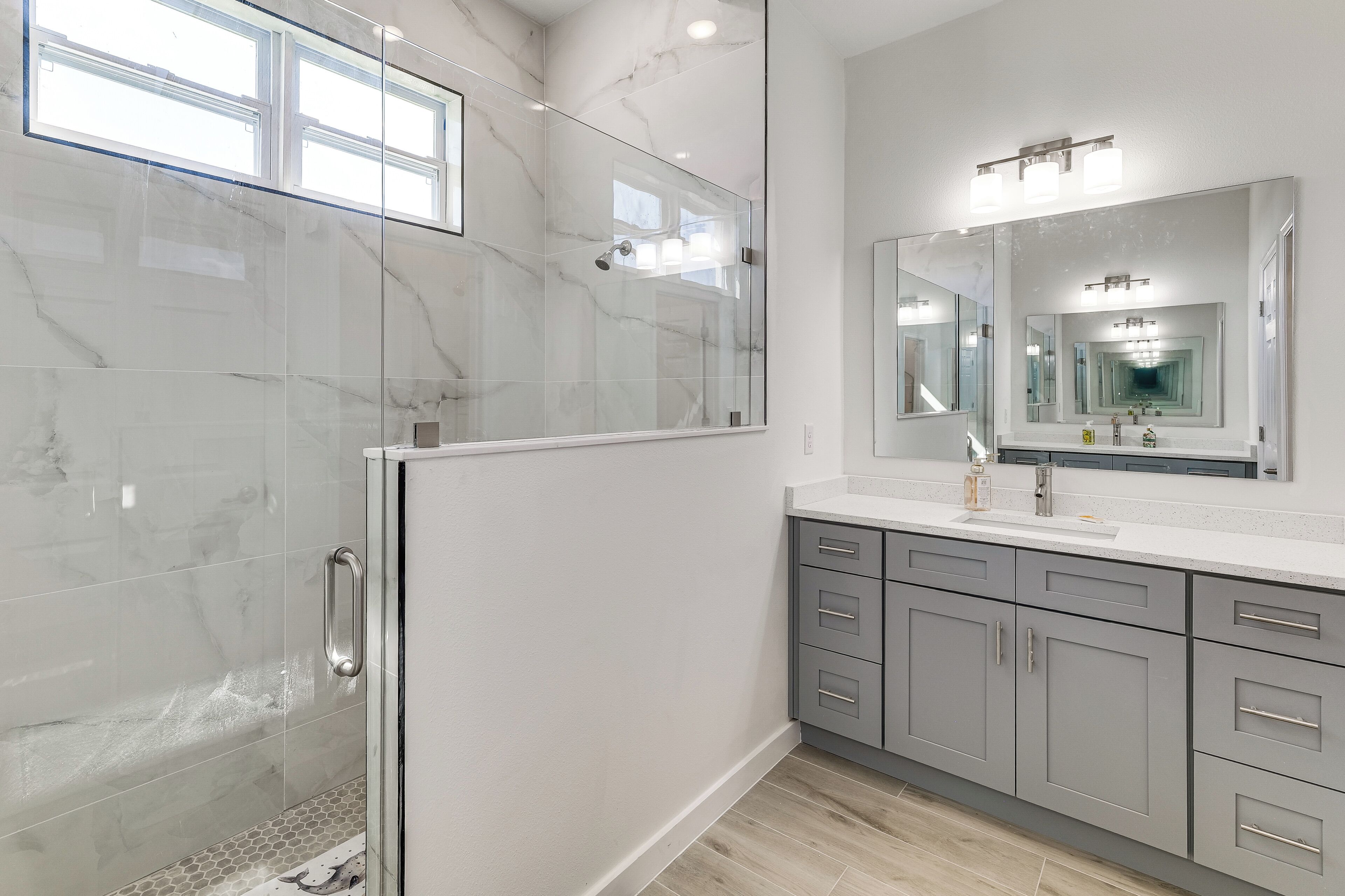A modern bathroom with a glass shower enclosure, a large mirror, and grey cabinets.