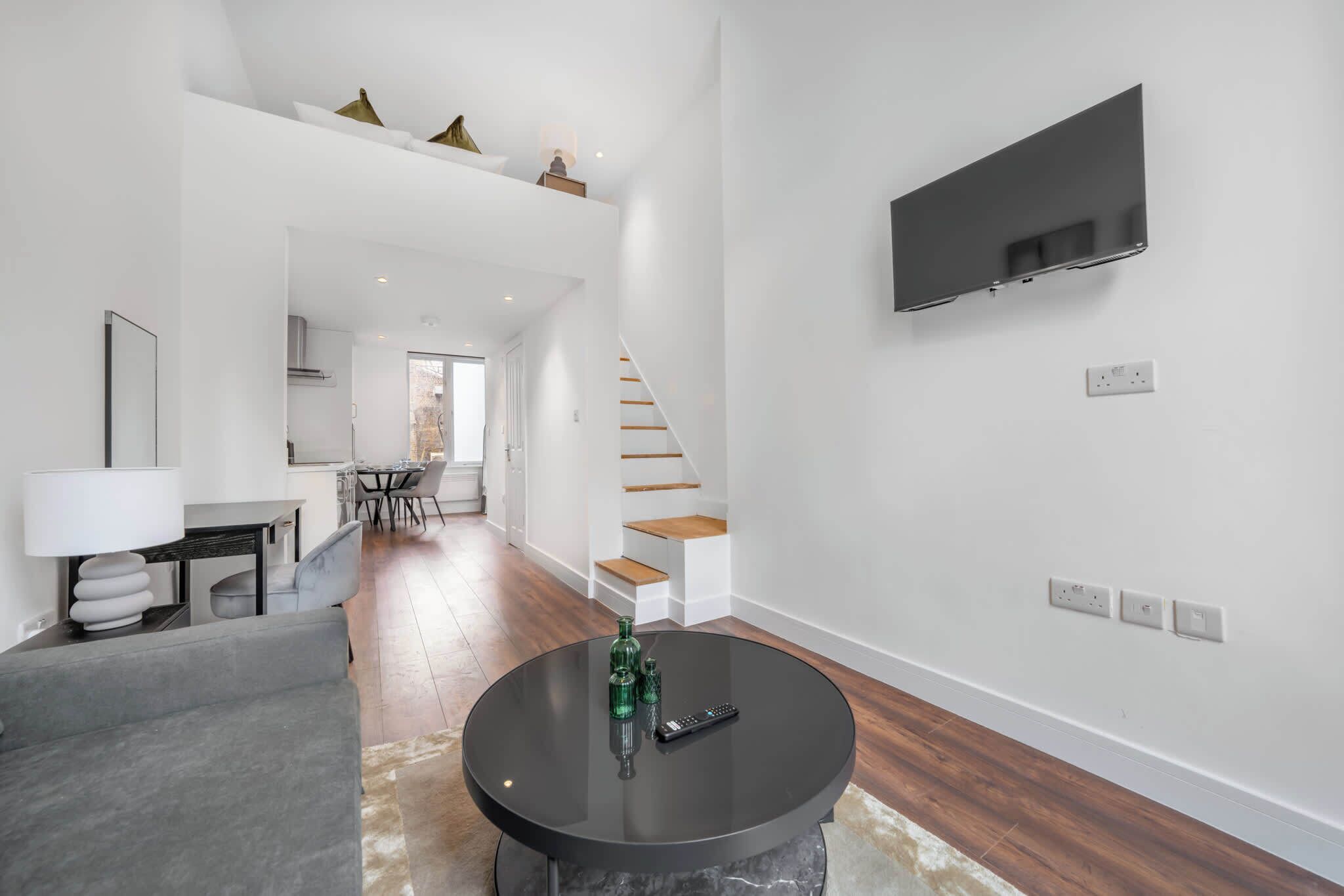 A modern living room with a flat-screen TV, a round coffee table with decorative items, a grey sofa, and a staircase leading to an upper level.