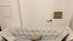 Ein helles, gepolstertes Sofa mit einem Zierkissen, zwei runde Holftische mit Drahtgestellen, eine Heizung und eine Tür mit kleinem Fenster.