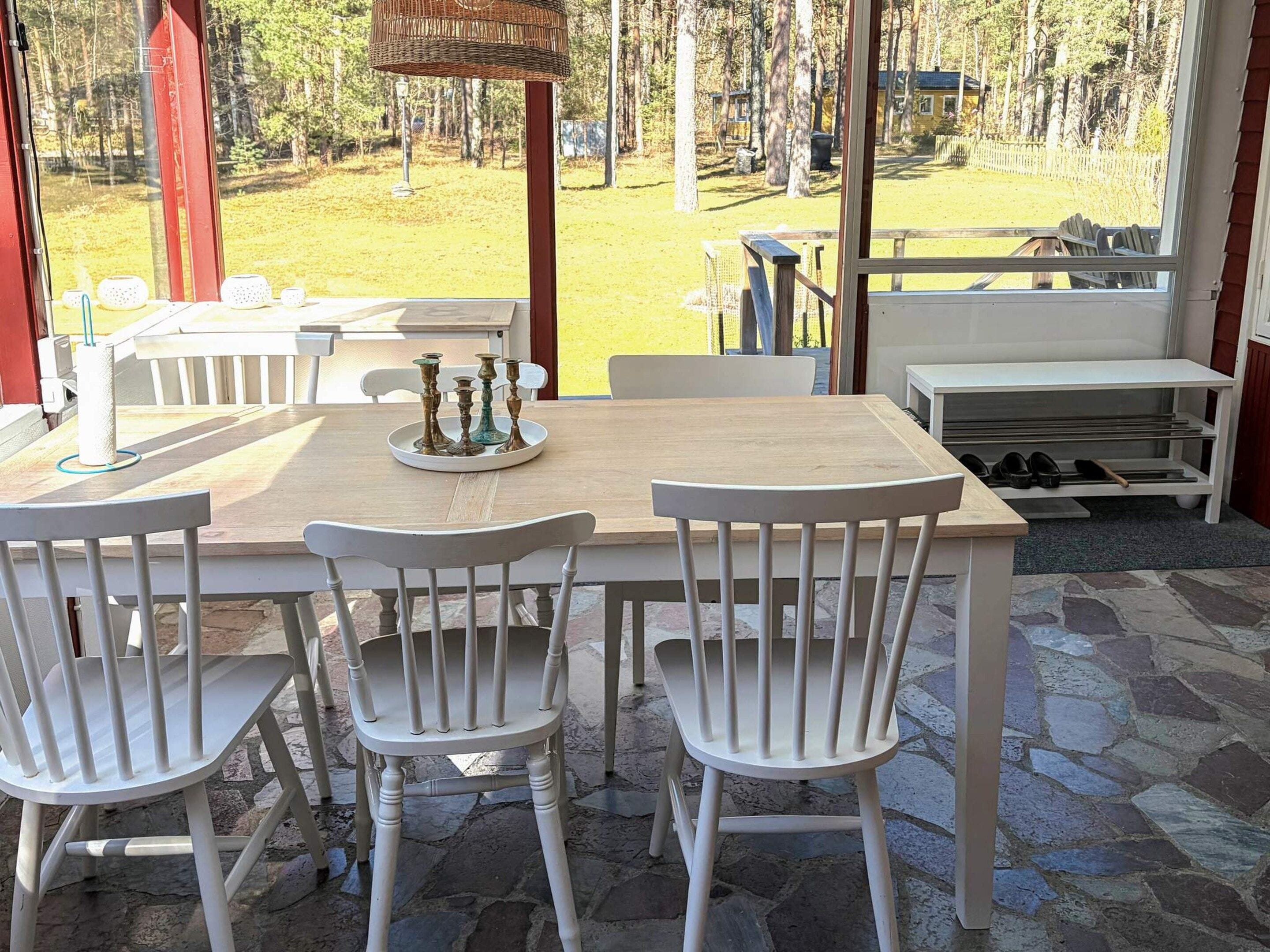 Un espacio exterior techado para comer con una mesa, sillas y una cesta de mimbre, con vistas a una zona boscosa.