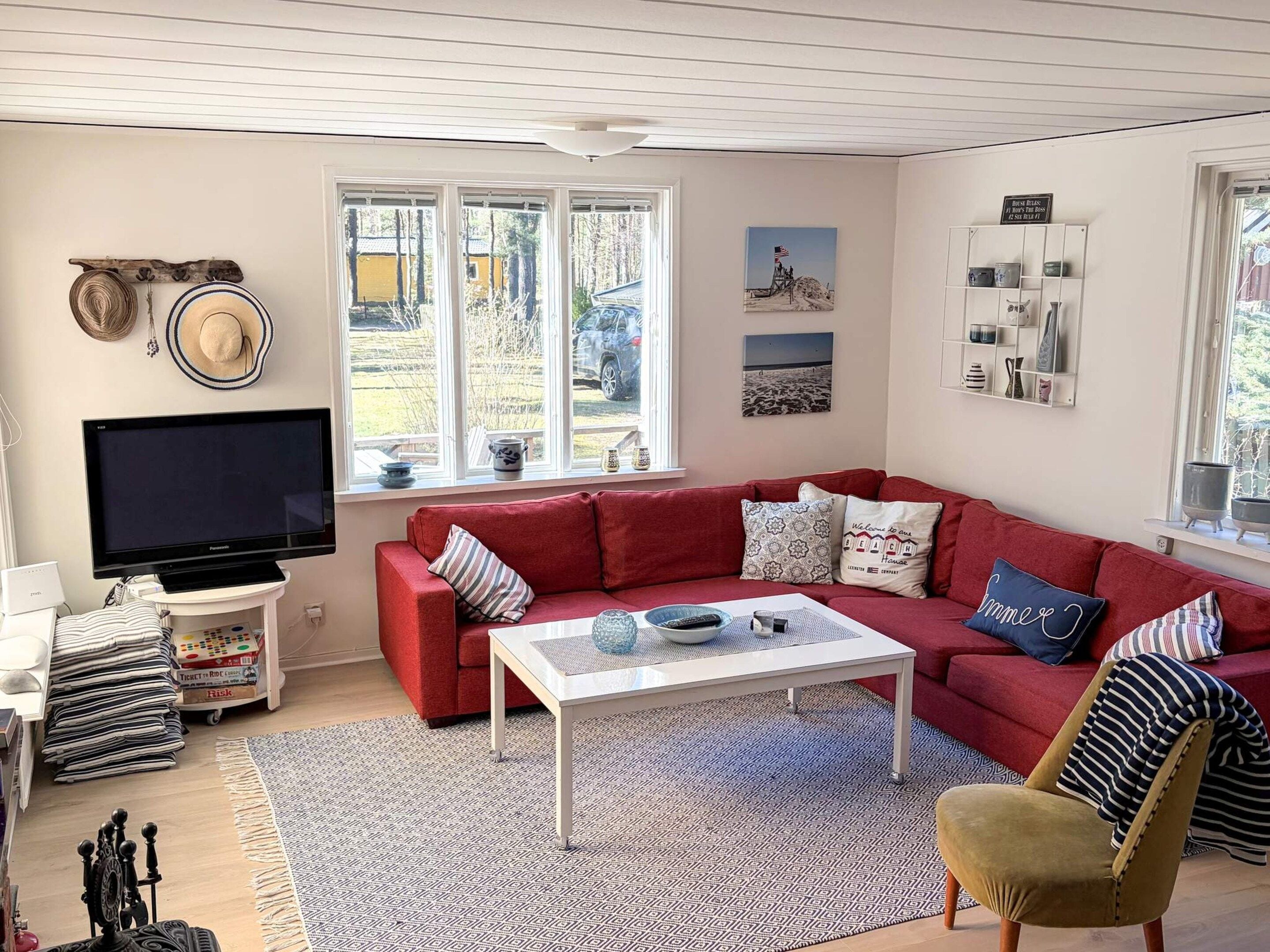Ett vardagsrum med en röd soffa, ett vitt soffbord och en tv. Det finns fönster med utsikt över omgivningen och en hylla med dekorativa föremål.
