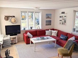 Ein Wohnzimmer mit einem roten Sofa, einem weißen Couchtisch und einem Fernseher. Es gibt Fenster mit Blick nach draußen und ein Regal mit Dekoration.