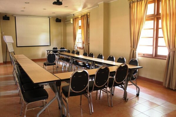 Un salón de conferencias con una mesa larga, sillas, una pantalla de proyector y ventanas con cortinas.