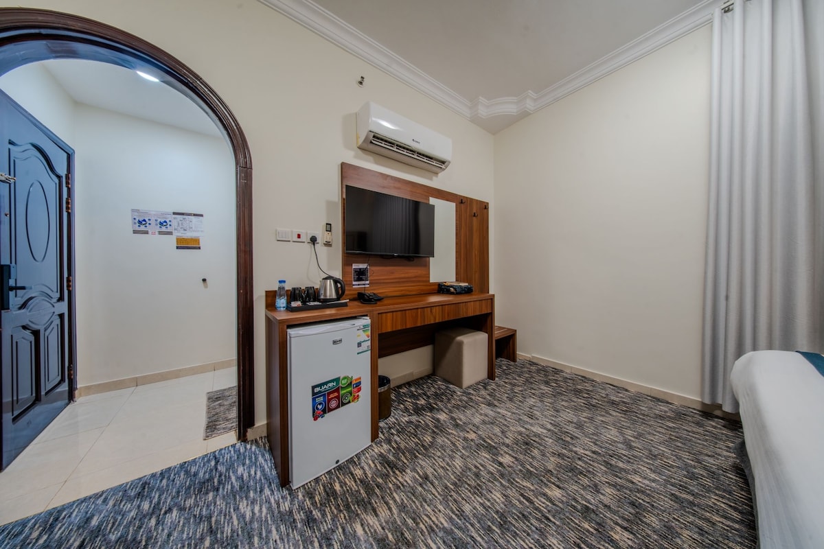 A hotel room with a bed, a desk with a mirror, a small refrigerator, an air conditioning unit, a television, and a door.