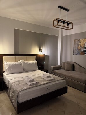 A modern bedroom with a large bed, a sofa, a nightstand, and a wall-mounted light fixture. - MG Luxury Living (Volvi)