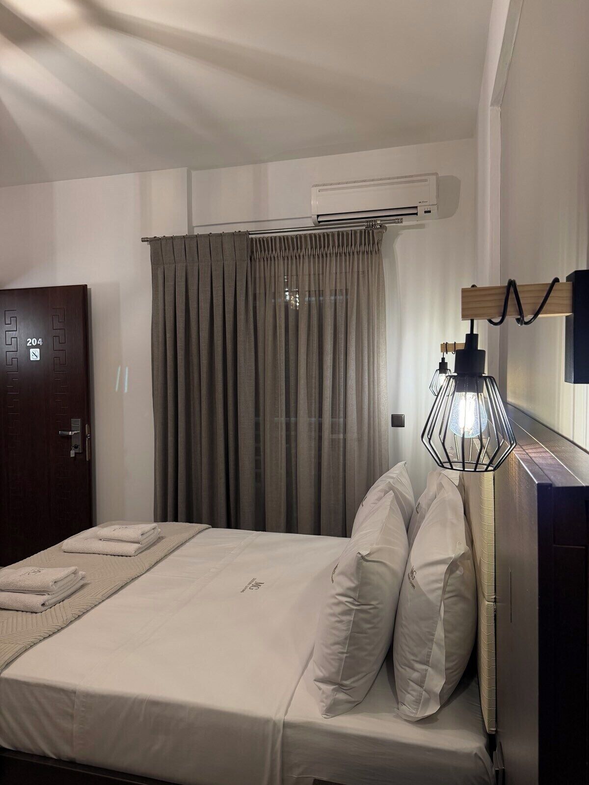 A hotel room with a bed, a wardrobe, a curtain, an air conditioning unit, and a lamp.