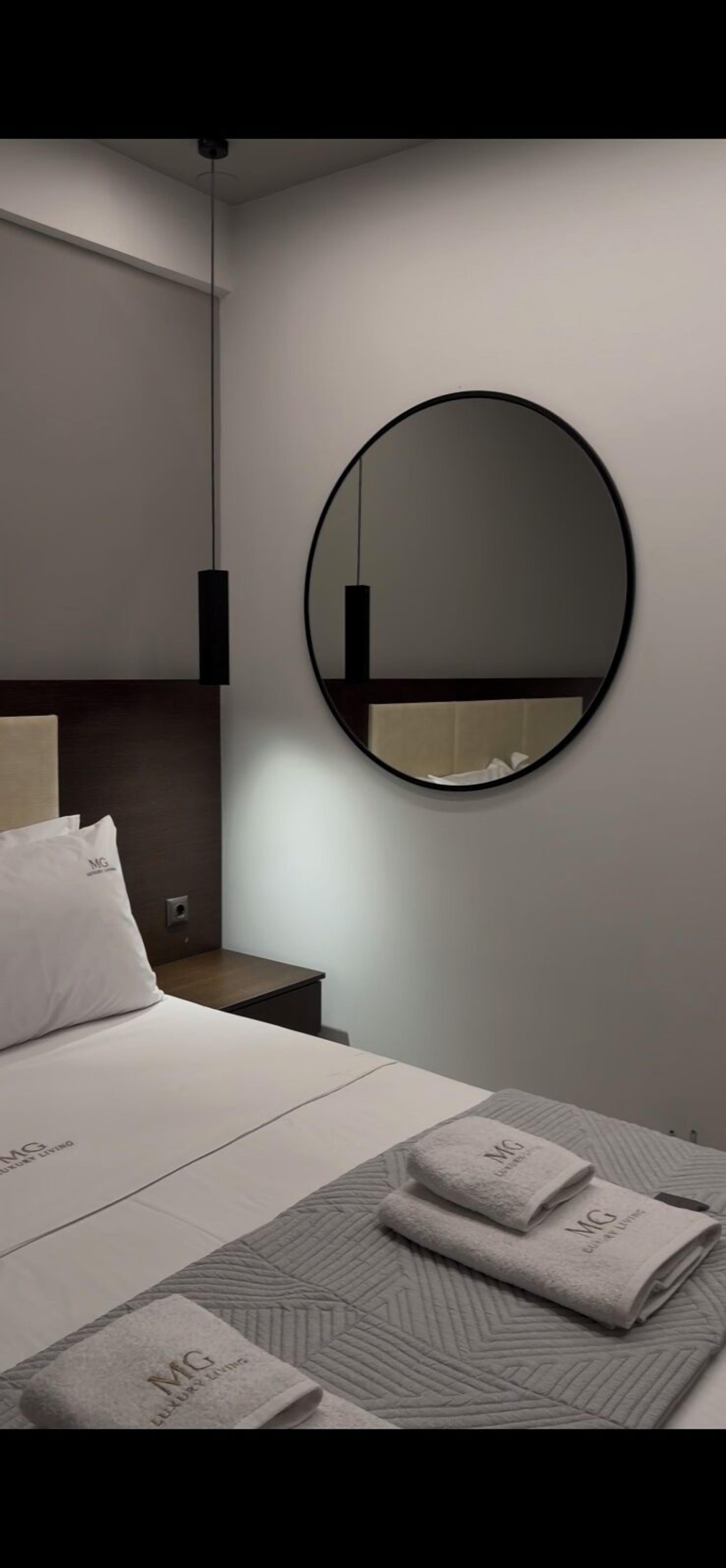 A modern hotel room with a bed, a round mirror, a nightstand, and neatly folded towels with 'MG' branding.