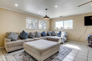 Una sala de estar con un sofá modular, reposapiés, alfombra estampada, lámpara de techo y ventanas amplias.