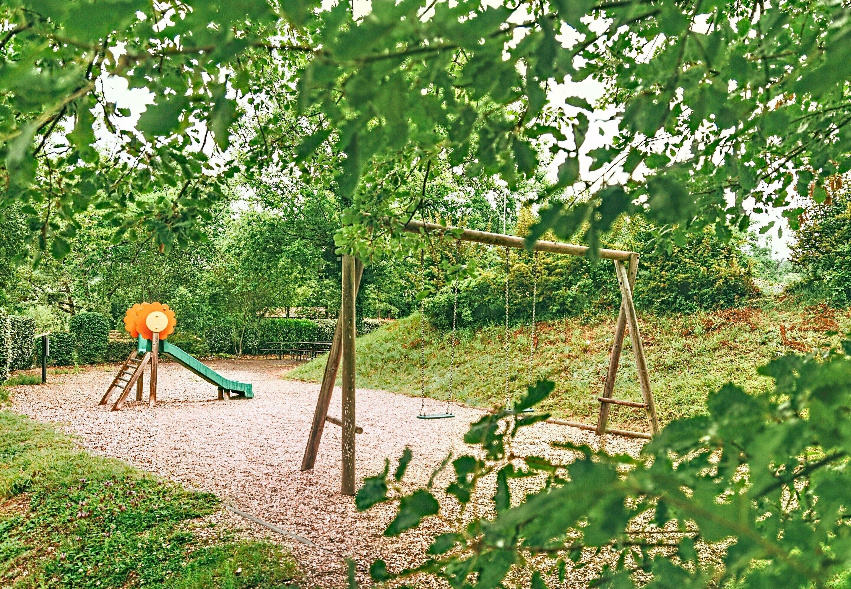 Un'area giochi con scivolo e altalene, circondata da alberi e un fondo in ghiaia.