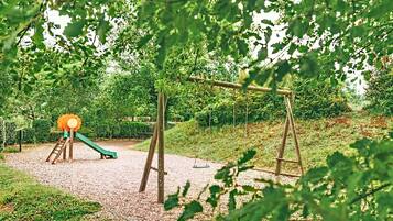 Un'area giochi con scivolo e altalene, circondata da alberi e un fondo in ghiaia.