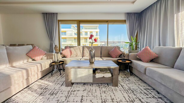 A modern living room with a sofa set, a coffee table, and a view of a cityscape through large windows. - The Carrousel Residence - Rabat (Rabat)
