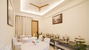 A hotel dining room with round tables, white chairs, a ceiling fan, and silver serving dishes on a shelf. - Treebo Premium Inn 62 (Noida)