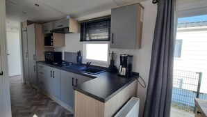 A compact kitchen with a microwave, stove, and sink, wooden cabinets, and a window with blinds. - Vakantiepark Kijkduin - 838 (Den Haag)
